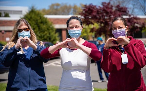 three people wearing surgical masks stand together making heart shapes with their hands