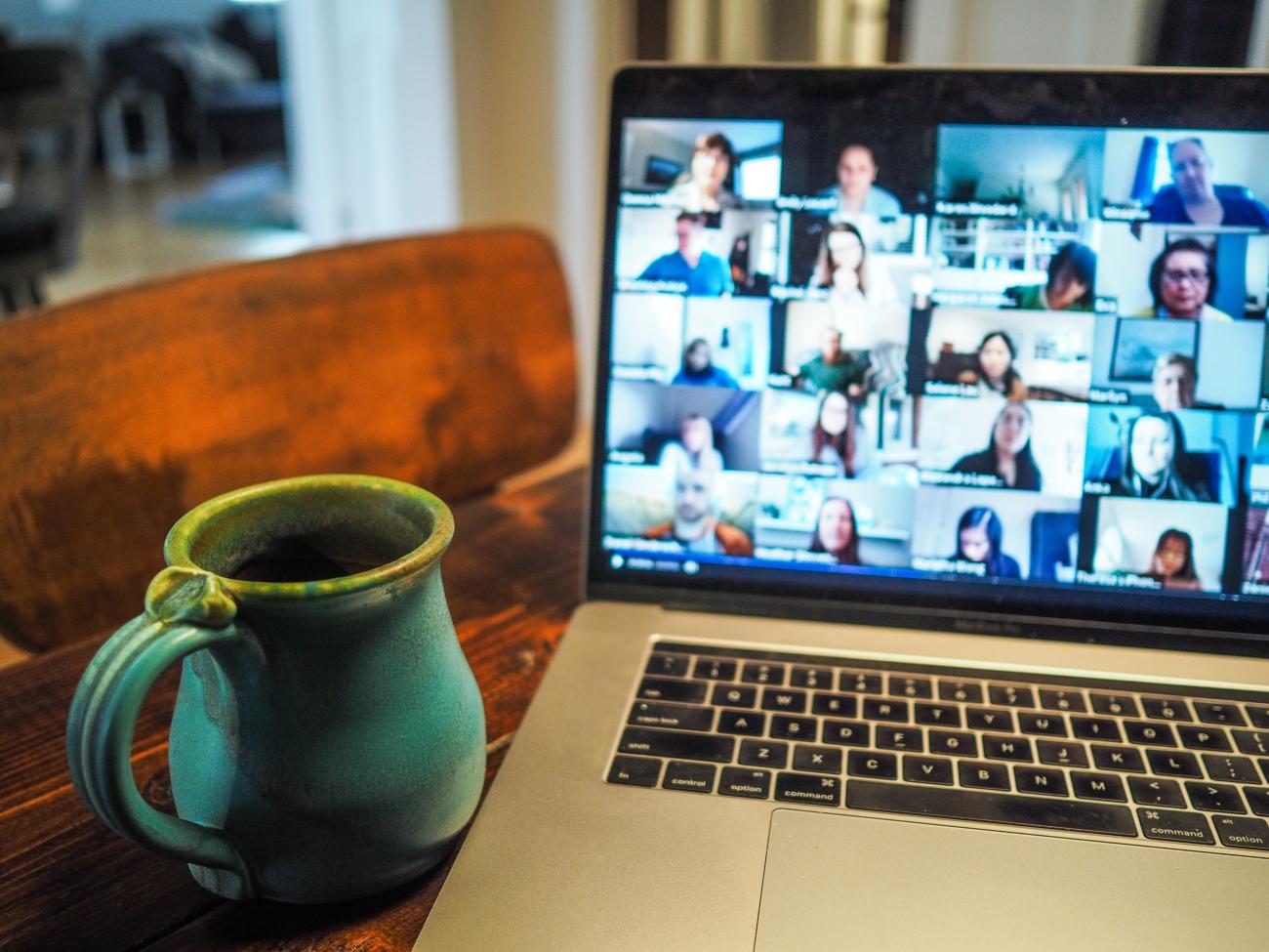 a laptop and mug on a living room desk from the point of view of someone on a video call. Various faces show on screen in a video conference layout.